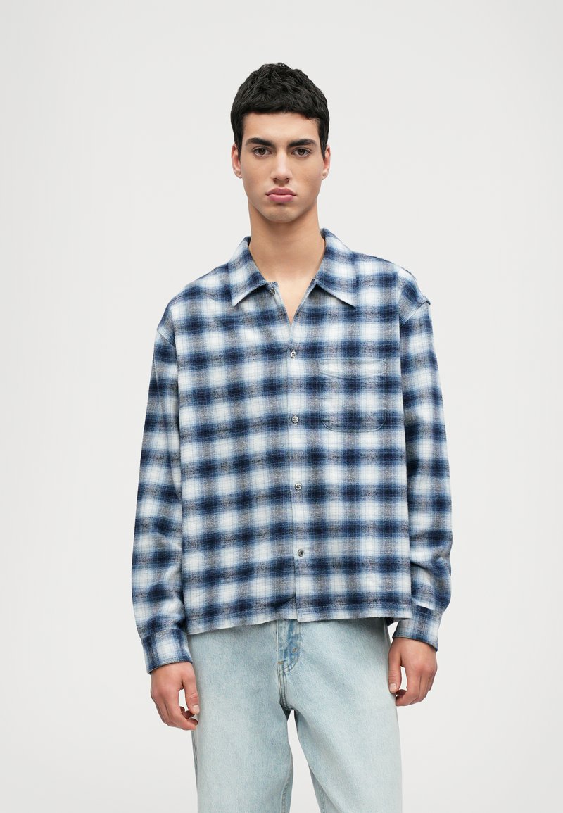 Καρό πουκάμισο σε μπλε και λευκές αποχρώσεις, φτιαγμένο από μαλακό ύφασμα, με κουμπιά μπροστά, μακριά μανίκια και τσέπη στο στήθος.