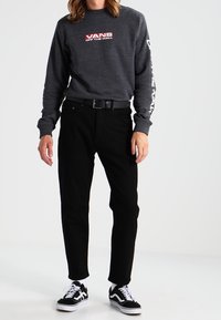 Harmaa collegepaita, jossa on "VANS OFF THE WALL" -logo, mustat farkut ja mustavalkoiset lenkkarit. Rentoon tyyliin sopiva pitkähihainen ja vyötäröltä istuva malli.