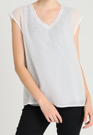 Vrouw draagt een losse, mouwloze witte blouse met V-hals, gecombineerd met een zwarte broek, staat met één arm licht gebogen.