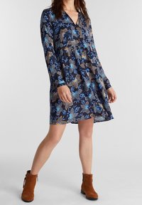 Abito blu navy con motivi floreali e a righe, scollo a V, maniche lunghe e una vestibilità rilassata. Indossato con stivaletti marroni alla caviglia.