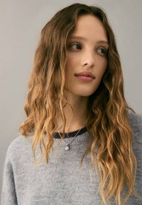 Osoba s dugom, valovitom kosom koja prelazi iz tamnosmeđe u svijetlosmeđu, nosi sivu, teksturiranu džemper i srebrnu ogrlicu s privjeskom.