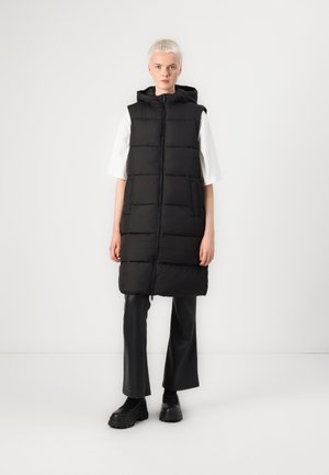Gilet rembourré noir avec une capuche, doté d'une fermeture éclair à l'avant, de deux poches latérales et d'une silhouette longue. Porté sur un haut blanc et un pantalon noir.