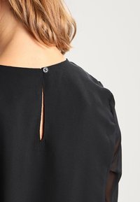 Schwarze Bluse mit einem Schlüssel-Loch-Ausschnitt im Rücken, ausgestattet mit einem kleinen Knopfverschluss und transparenten Ärmeln. Glatte Materialbeschaffenheit mit einem minimalistischen Design.