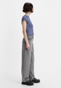 Šedé džínsy s širokými nohavicemi v kombinaci s krátkým topem v levandulové barvě s vykrojenými okraji. Outfit doplňují černé platformové boty.
