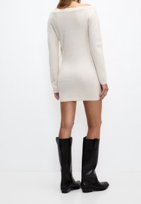 Robe en maille crème à manches longues avec un design épaules dénudées, tissu texturé et silhouette ajustée, associée à des bottes en cuir noires montantes jusqu'au genou.