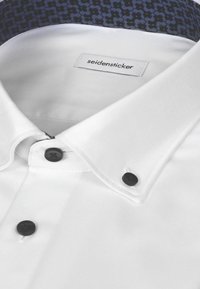 Chemise blanche avec boutons noirs et une étiquette à l’intérieur du col indiquant "seidensticker," avec une doublure intérieure à motifs.
