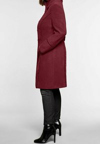 Abrigo de lana burdeos con cuello alto y corte entallado. Presenta costuras laterales y bolsillos. Lleva puesto sobre unos jeans grises y botas de tacón negro.
