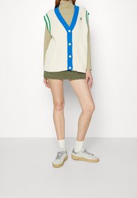 Ljusbeige stickad väst med blå knappar och grön kant, lagd över en olivgrön polotröja. I kombination med olivgröna shorts och ljusgrå sneakers.