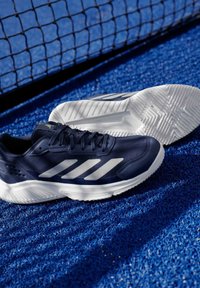 Двойка маратонки Adidas в тъмносиньо и бяло на синя текстурирана спортна площадка близо до тенис мрежа.