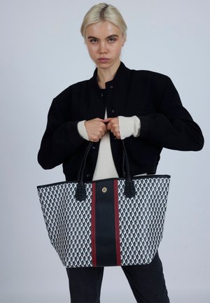 Velika torba za nošenje izrađena od uzorkovanog tkanog materijala u mornarsko plavoj i bijeloj boji, središnjim crvenim prugom i crnim detaljima; glatka tekstura s čvrstim ručkama.