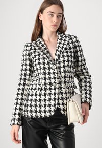 Sivi i bijeli blejzer u houndstooth uzorku s teksturiranim materijalom, s jednim redom dugmadi, rever kragnom i zatvaranjem na dva dugmeta.