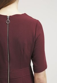 Vestido burdeos con mangas cortas y silueta ajustada, con un cierre metálico visible en la parte posterior y una costura que acentúa la cintura.