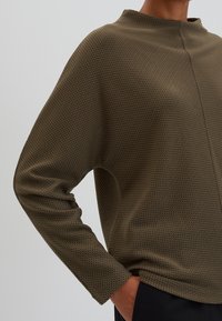 Hnědý pulovr se vzorem, vysokým límcem, dlouhými rukávy a volným střihem. Materiál má měkkou texturu a jemný geometrický design.