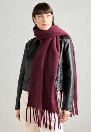 Écharpe en laine bordeaux avec des franges, portée autour du cou, associée à une veste en cuir noir et un pantalon clair.