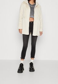 Abrigo de piel sintética de color crema con capucha, combinado con un top negro corto, jeans negros desgastados con franjas laterales blancas y botas negras gruesas.