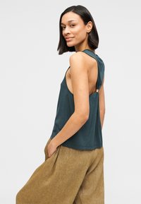 Top sin mangas color azul verdoso con un ajuste relajado, diseño de espalda abierta y un detalle dorado en el hombro, combinado con pantalones marrones de pierna ancha.