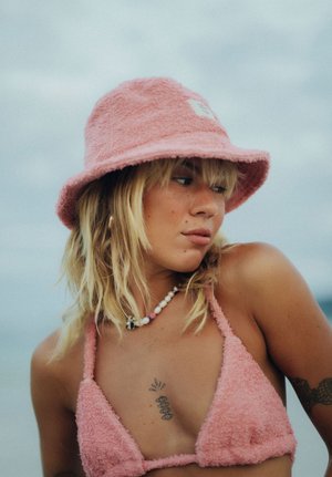 Młoda kobieta o blond włosach, ubrana w różowy, puszysty kapelusz typu bucket oraz pasujący top bikini, z naszyjnikiem z koralików i tatuażami na klatce piersiowej oraz ramieniu.