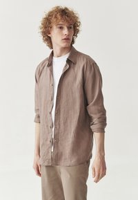 Cămașă button-up de culoare maro deschis, dintr-un material texturat, cu guler, mâneci răsucite și tiv drept. Purtată peste un tricou alb.