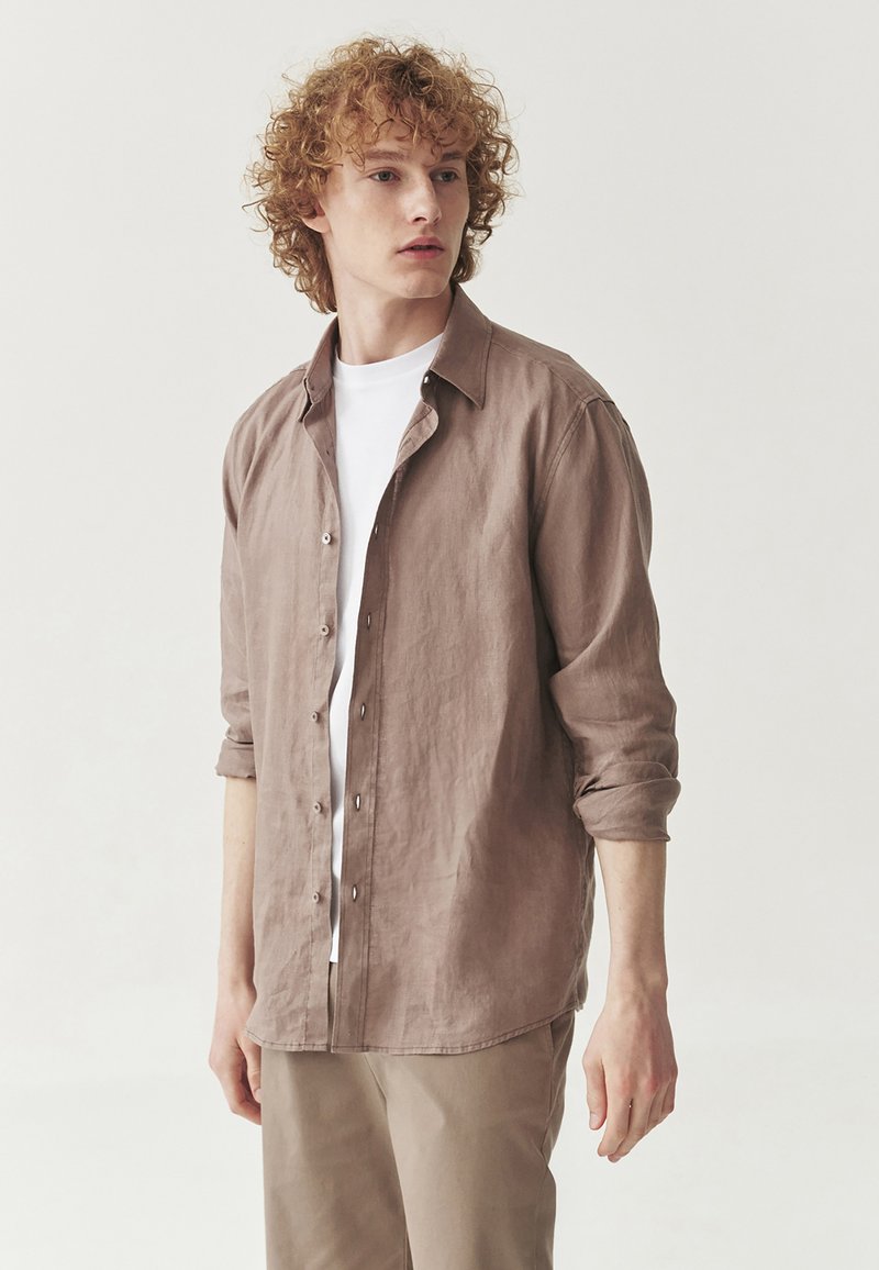 Cămașă button-up de culoare maro deschis, dintr-un material texturat, cu guler, mâneci răsucite și tiv drept. Purtată peste un tricou alb.