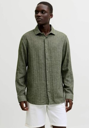 Mann trägt ein olivgrünes Hemd mit langen Ärmeln und weiße Shorts, steht vor einem schlichten hellgrauen Hintergrund und schaut nach links.