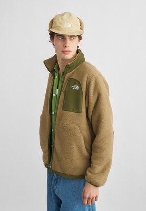 Junger Mann trägt eine beige Fleecejacke mit olivgrünen Akzenten, grünes Hemd, blaue Jeans und beige Mütze mit Ohrklappen, schaut nach links.