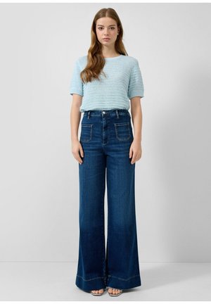 Młoda kobieta z długimi włosami stoi, ubrana w jasnoniebieską, krótkorękawową dzianinową bluzkę oraz granatowe, szerokie spodnie z wysokim stanem i przednimi kieszeniami naszywanymi.