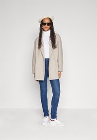 Beige Oversize-Mantel, weißer Rollkragenpullover, blaue Skinny Jeans, weiße Sneaker mit roten Akzenten, cremefarbene Beret, dunkle Sonnenbrille. Schlichtes Design, lässiger Look.