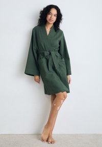 Robe en kimono vert texturé avec des manches longues, une taille ceinturée et des poches latérales ; le mannequin se tient pieds nus contre un fond neutre.