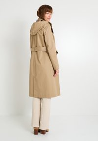 Trench coat de color beige claro con capucha, cinturón de cintura ajustable y mangas largas. Textura suave, de longitud a la rodilla y con un diseño sencillo.