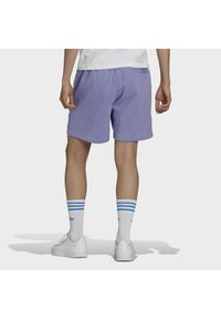 Muž v fialových šortkách, bílých polokozačkách Adidas s modrými pruhy a bílých teniskách, stojící na pozadí s jednoduchým vzorem čelem k divákovi.