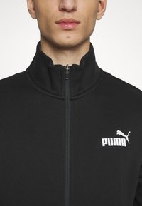 Black zip-up jacket with high collar, featuring a smooth texture and a white Puma logo on the left chest. Zipper in the center front.