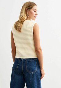 Strickärmloses Oberteil in hellem Beige mit einem strukturierten, gerippten Muster. Kombiniert mit hochgeschnittenen, dunkelblauen Jeanshosen mit Gesäßtaschen.