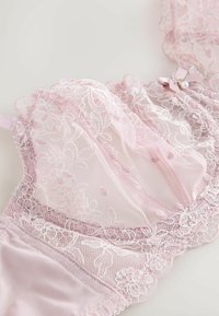 Delikatny jasnoróżowy koronkowy biustonosz z kwiatowym haftem, przejrzystymi miseczkami z siateczki, falistymi krawędziami oraz małą kokardką z perłowym zawieszką na białym tle.