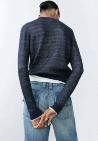 Marineblauwe gebreide trui met lange mouwen, voorzien van een geribde zoom. Draagt men met lichtblauwe denim jeans met een losse pasvorm.