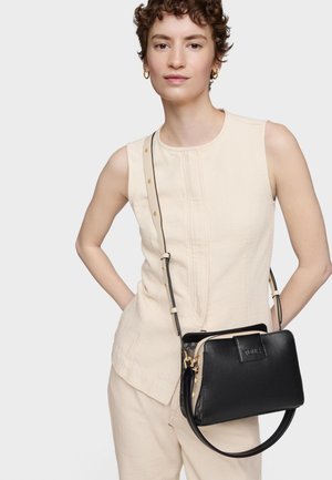 Donna che indossa un abito crema senza maniche e orecchini a cerchio d'oro, con una borsa a tracolla in pelle nera con dettagli in oro.