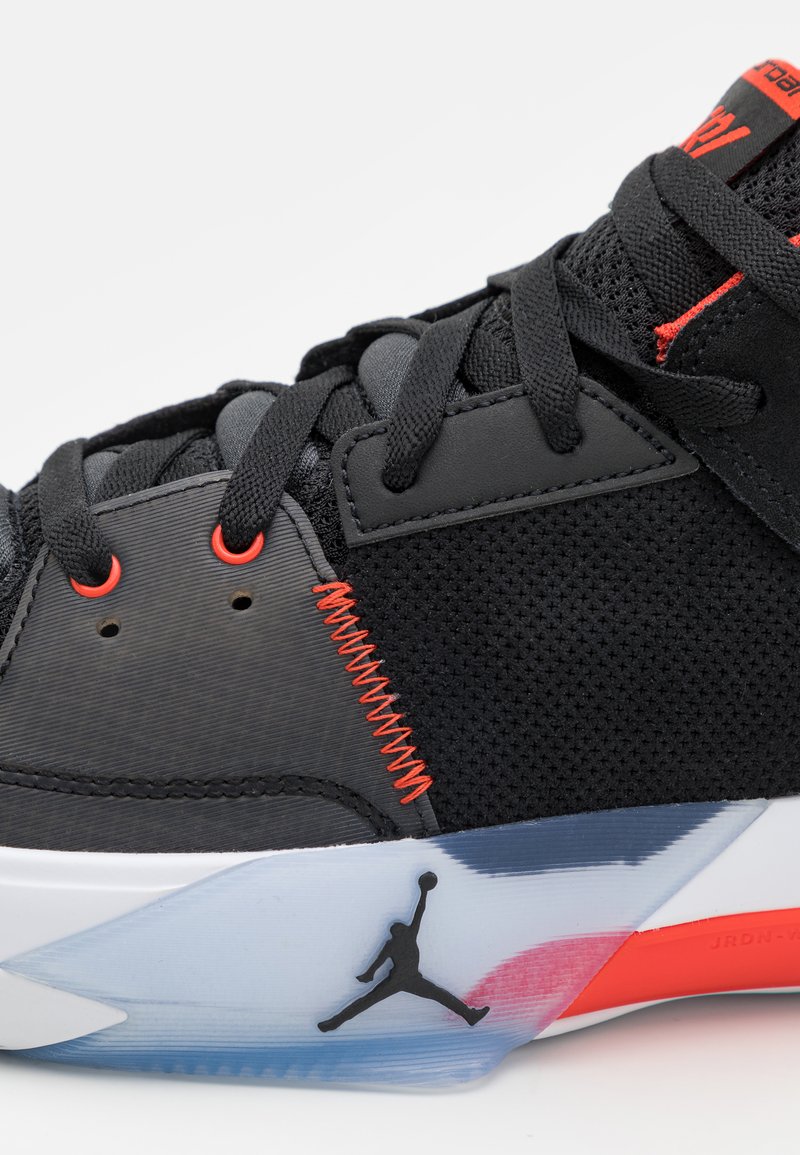 Chaussure de sport noire en mesh et matériaux synthétiques, avec des accents rouges vifs, des motifs texturés et une semelle translucide ornée d'un détail de logo.
