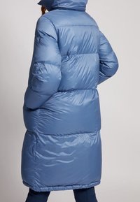 Veste d'hiver bleue longue et rembourrée avec une capuche, portée par une personne de dos les mains dans les poches sur un fond uni.