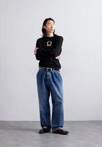 Schwarzer Pullover mit goldenem Logo, lässige blaue Jeans im relaxed Fit und schwarzen Schuhen. Schlichtes Design und lässige Passform mit strukturierter Oberfläche.