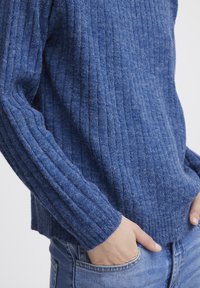 Sinine soonikkoes, mis on valmistatud pehmest kangast, koos pikkade varrukatega ja ümmarguse äärega, sobitub denimpükstega. Tekstuur tundub hubane ja soe.