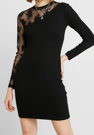 Kvinde iført en tætsiddende sort kjole med én gennemsigtig ærme i blomstret blonder og lagdelte halskæder mod en ensfarvet baggrund.