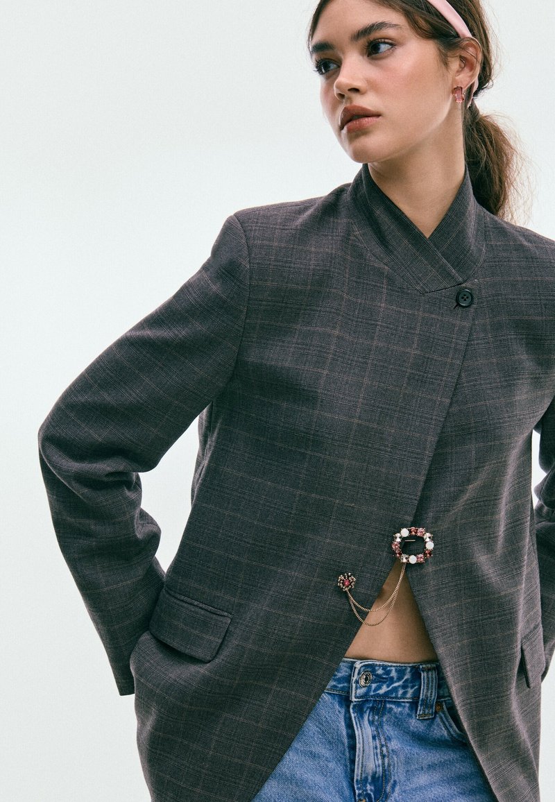 Jeune femme portant un blazer à carreaux gris avec des broches décoratives, un jean bleu et un bandeau rose, regardant sur le côté sur un fond uni.