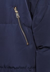 Námornícky pufovaný kabát s prostorem na zip a zlatými detaily, hladkou texturou a prošívaným designem, který zdůrazňuje teplo a funkčnost.