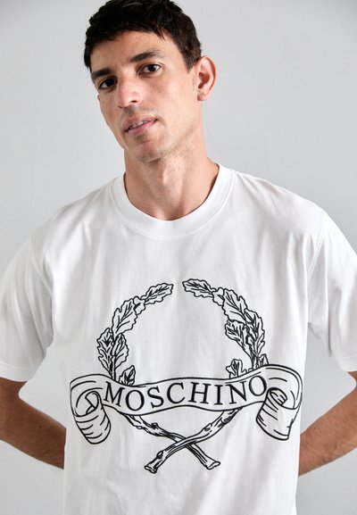 Bílá bavlněná tričko s černým grafickým designem vavřínových listů a stuhy s nápisem "MOSCHINO" uprostřed.