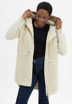 Person adjusting the hood of a cream-colored fleece-lined jacket over a black top and dark blue jeans against a plain background.