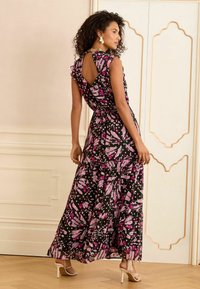 Fekete anyagú maxi ruha élénk rózsaszín és lila virágmintával, fodros ujjakkal és nyitott hátrésszel. Fehér sarokcipőkkel viselve.