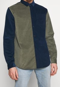 Nappipaita, jossa kaksivärinen muotoilu: toisella puolella oliivinvihreää samettikangasta ja toisella puolella laivastonsinistä, jossa on rintatasku ja napit.