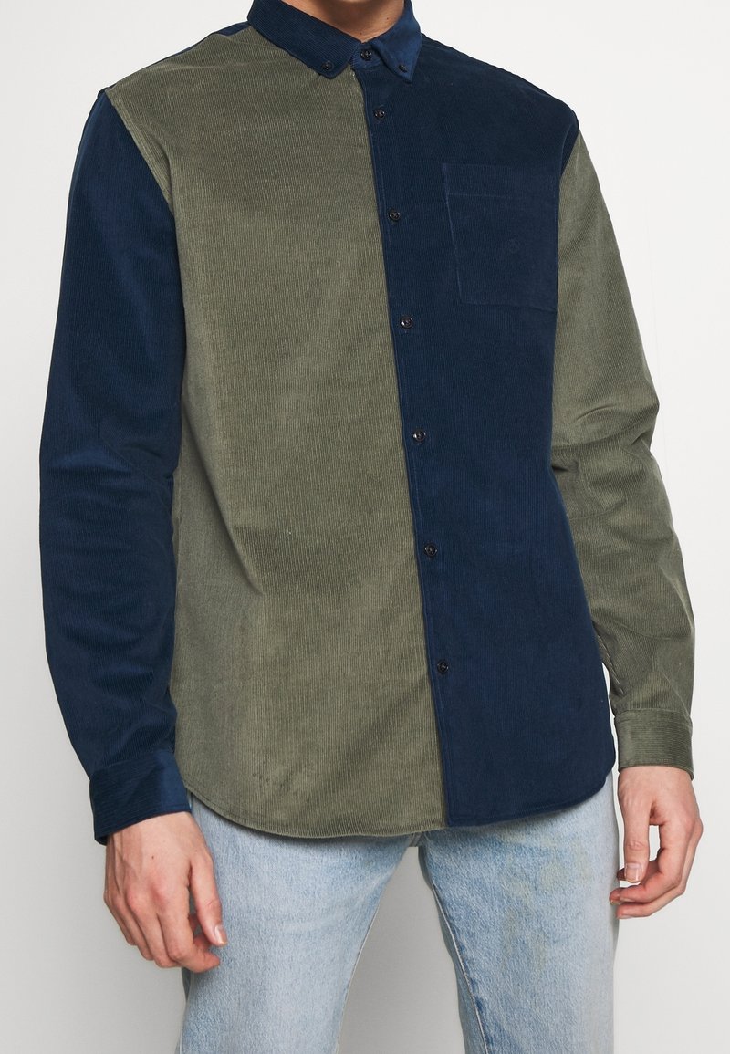 Nappipaita, jossa kaksivärinen muotoilu: toisella puolella oliivinvihreää samettikangasta ja toisella puolella laivastonsinistä, jossa on rintatasku ja napit.