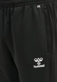 Primo piano di pantaloni della tuta neri con vita elastica, tasche laterali, logo Hummel bianco e iniziali XK vicino alla tasca.