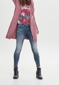 Langer pinker Cardigan über einem floral gemusterten weinroten Shirt, kombiniert mit hellblauen, zerstörten Skinny Jeans und schwarzen Ankle Boots.
