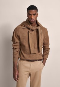Brauner Strickpullover mit strukturiertem Muster, kombiniert mit einem braunen Hemd mit Kragen darunter. Helle beigen Hosen und ein brauner Gürtel runden den Look ab.